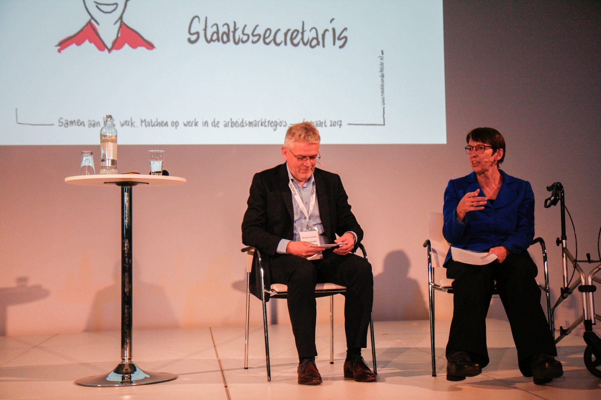 Jetta Klijnsma et al. zittend in stoelen voor een groot scherm