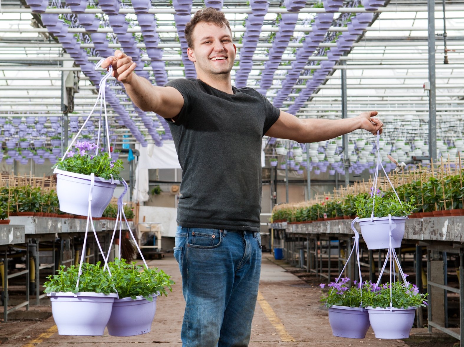 Pantar-medewerker Jerry houdt met een vrolijk gezicht 3 planten in hangpotten vast in een kas.