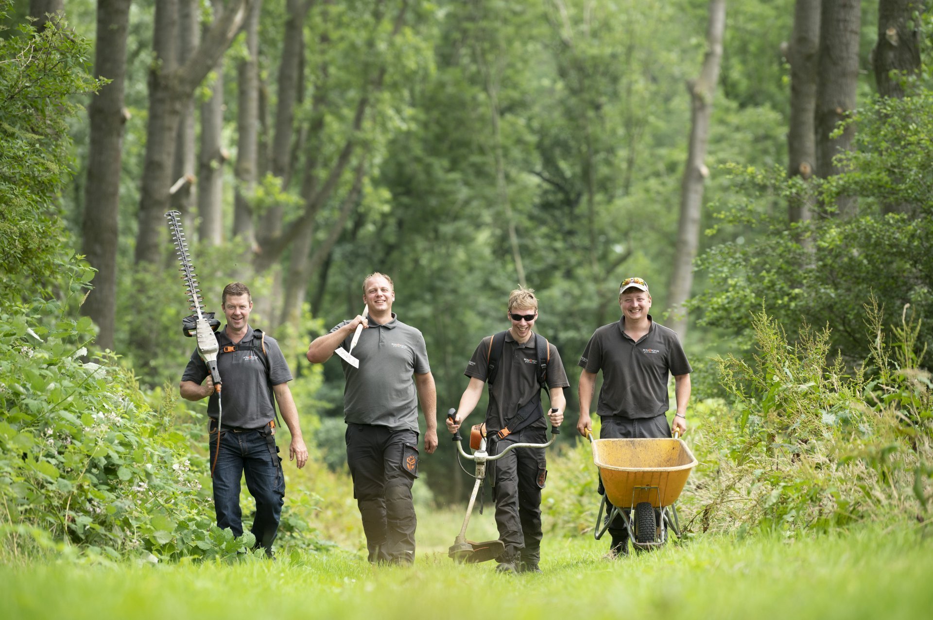 4 lachende mannen in werkkleding van Rosa Novum en met een heggenschaar, schoffel, bosmaaier en kruiwagen lopen in een bos