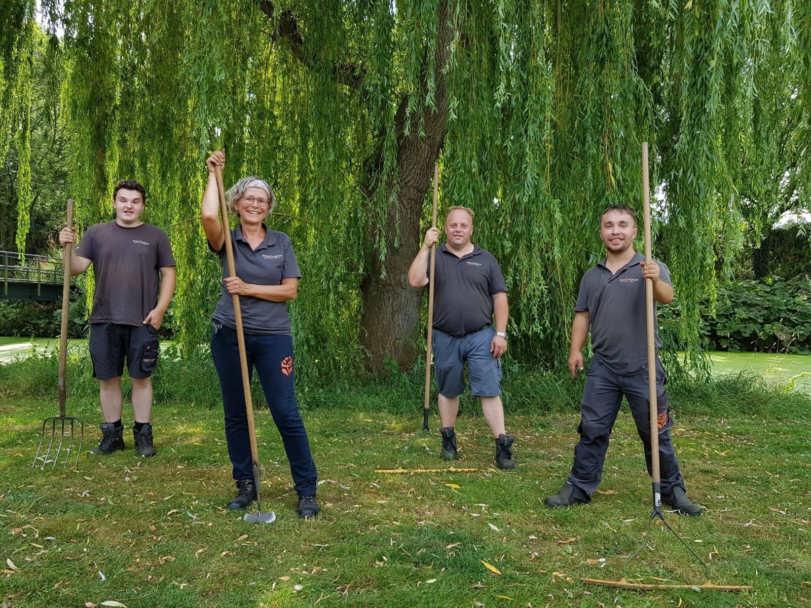 Nancy van der Vin (met schoffel) staat met 3 groenmedewerkers met spitvork en grasharken voor een treurwilg in het gras. van Rosa Novum,