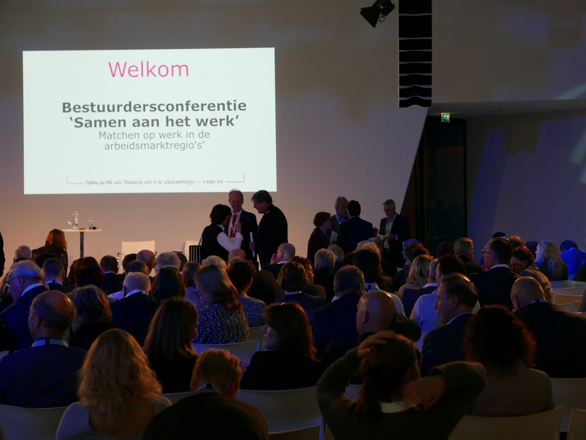 een groep mensen die in een ruimte met een projectiescherm zitten