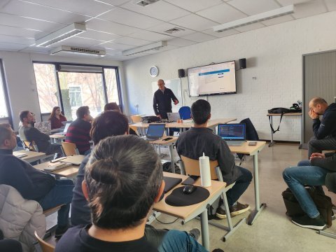 Ongeveer 10 leerlingen met laptops voor zich zitten in een klaslokaal. Voor de klas staat een docent voor een whitebord iets te vertellen.